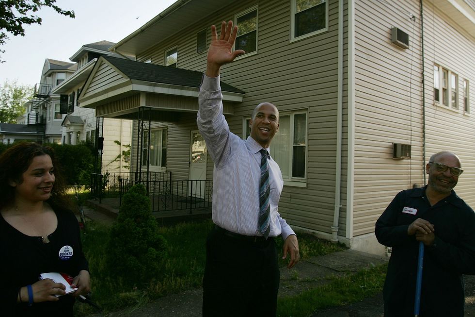 a woman and two men stand outside a house smiling, the man in the middle waves