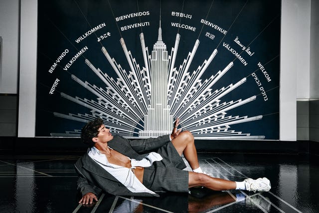 person lounged in a stylish gray suit against a wall with multiple welcome messages in different languages and an iconic building illustration