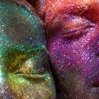 two faces, both covered in glitter, are cheek to cheek, eyes closed one is covered in blue and green glitter, the other in pink and purple glitter