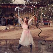 actress madelaine petsch playing with a water hose and looking happy, holding hose up over head with both hands while water pours out of hose, wearing black boots and a pink bralette and bottoms underneath a semi sheer white dress, in front of a farm background