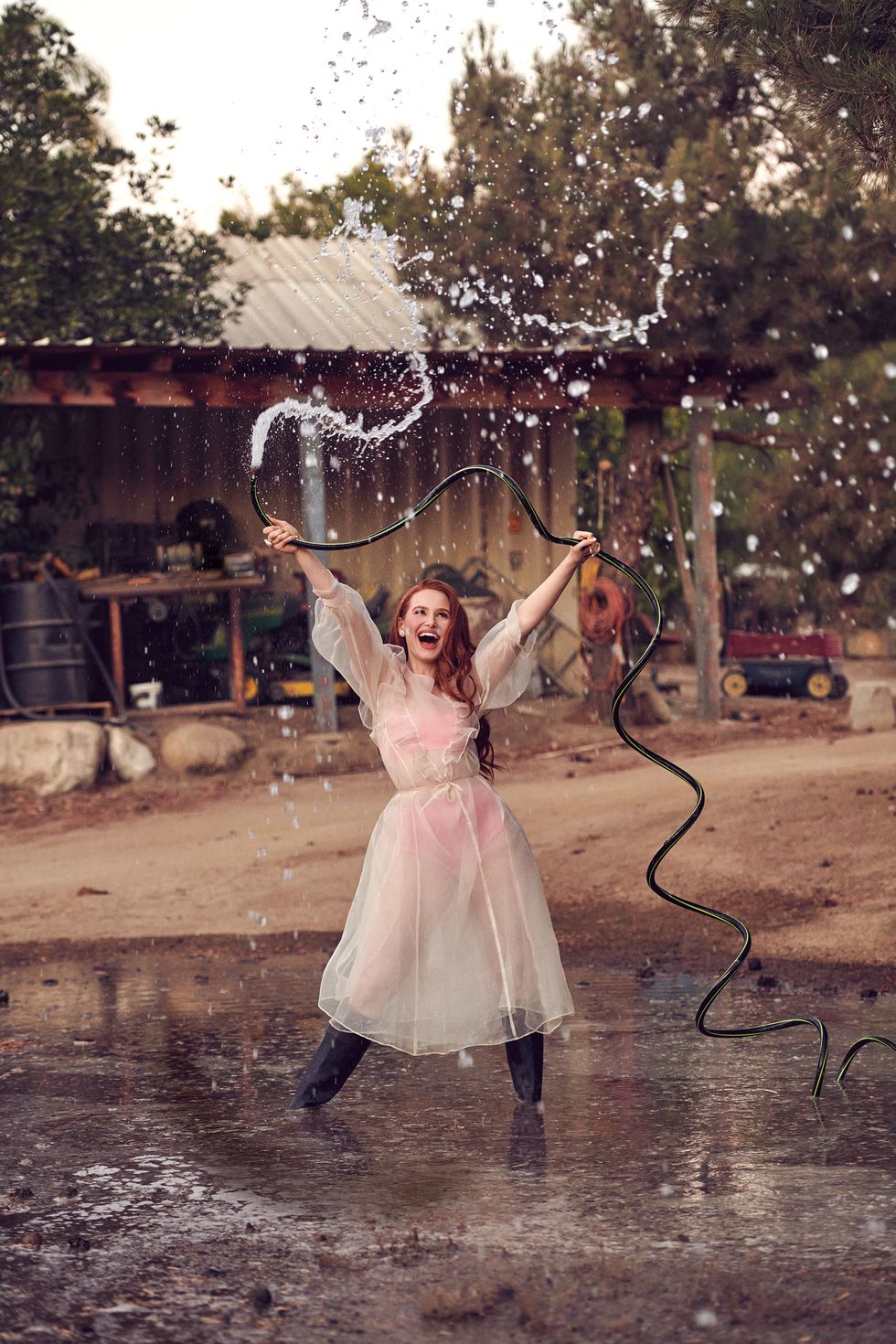 actress madelaine petsch playing with a water hose and looking happy, holding hose up over head with both hands while water pours out of hose, wearing black boots and a pink bralette and bottoms underneath a semi sheer white dress, in front of a farm background