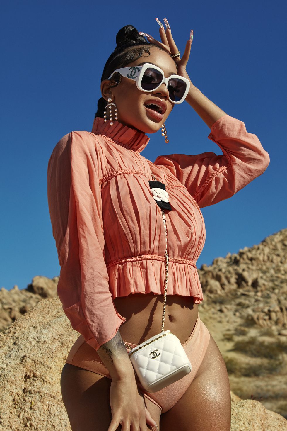 singer saweetie standing up with hand on head, wearing a salmon pink top and boyshorts with white sunglasses and a white clutch, in front of a desert and sky background