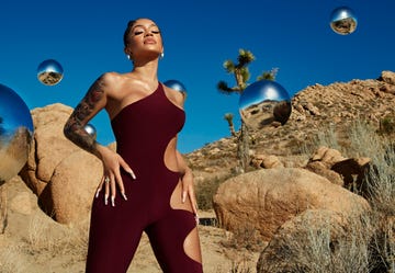 singer saweetie standing up with hands on hips and eyes closed, wearing a plum or wine colored jumpsuit, in front of a sky and desert background with reflective silver globes