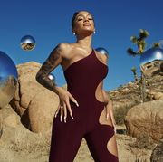singer saweetie standing up with hands on hips and eyes closed, wearing a plum or wine colored jumpsuit, in front of a sky and desert background with reflective silver globes