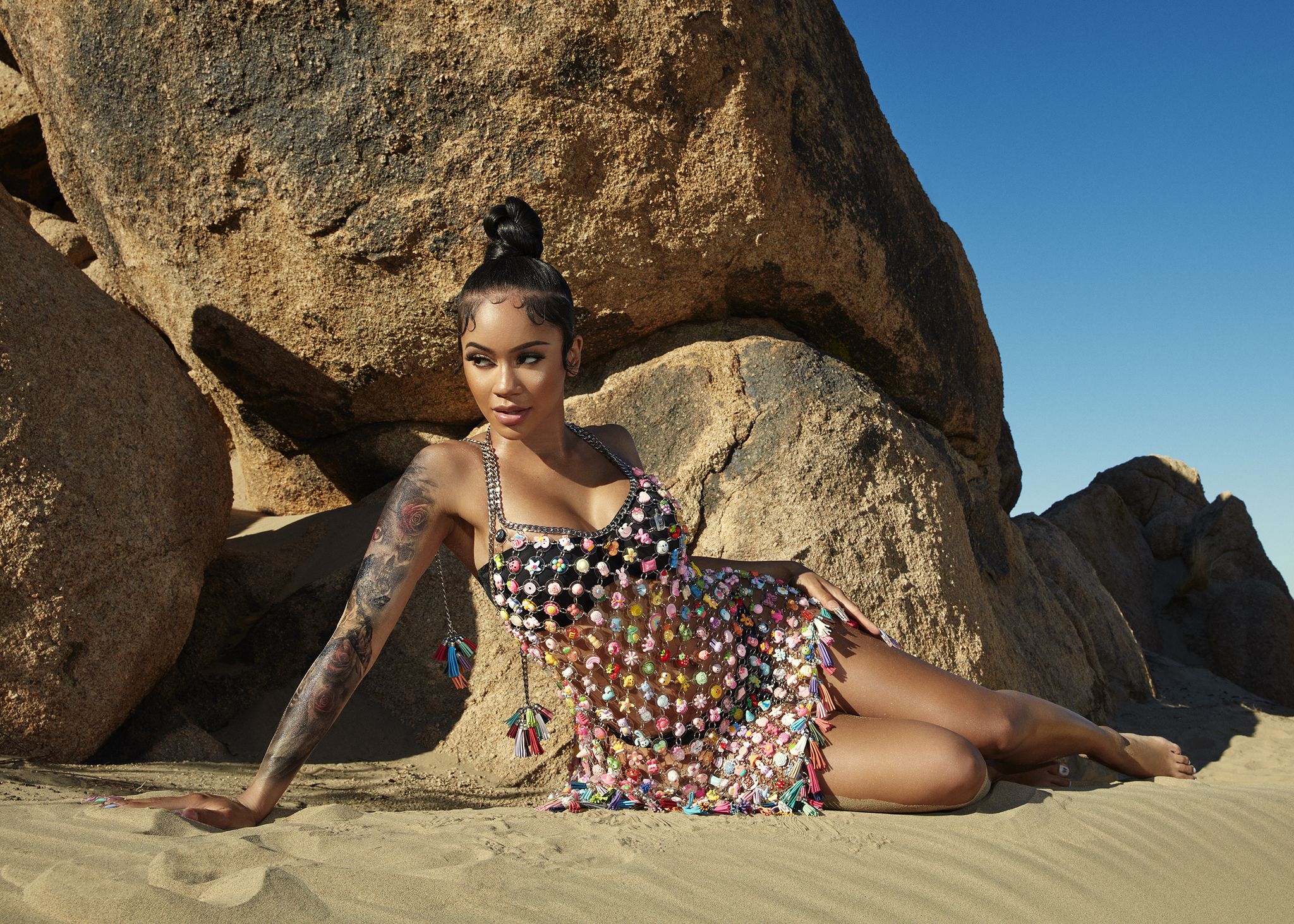 singer saweetie propped up with arm lying down on sand, wearing a colorful chain dress, in front of a desert and sky background