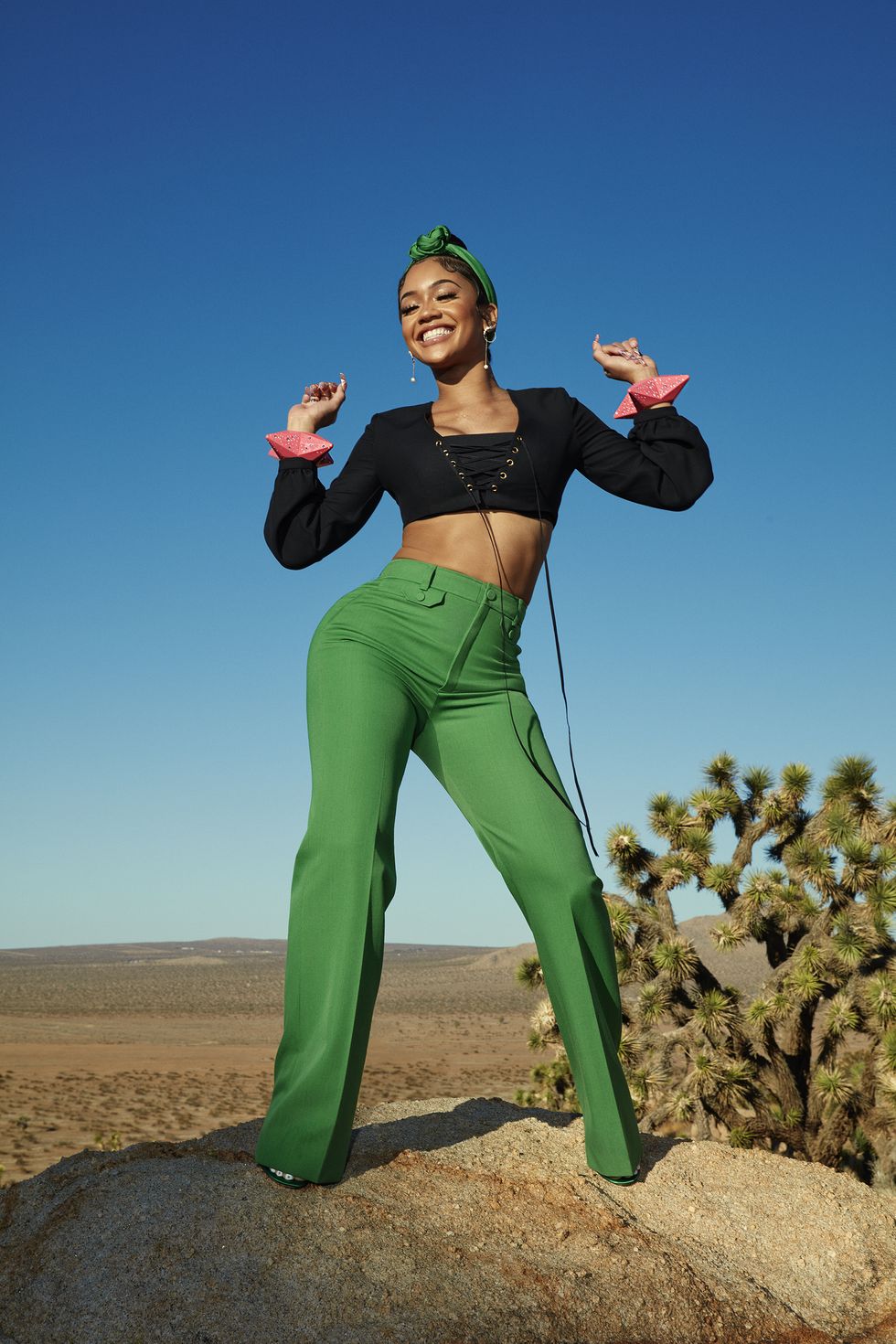 singer saweetie smiling and standing up with hands up in the air near each of her shoulders, wearing a black top, green pants, green headwear, and green heels, in front of a desert and sky background
