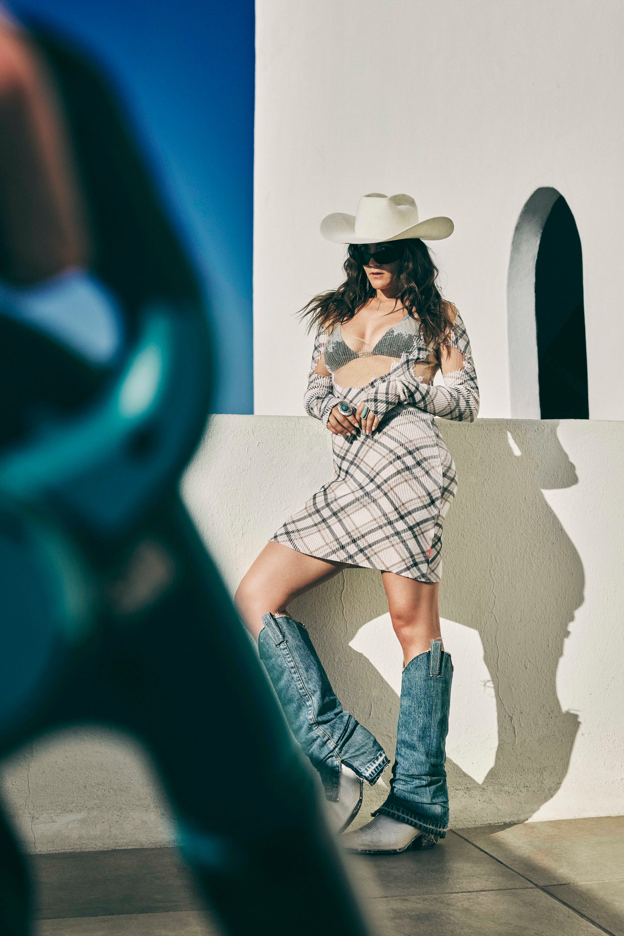 woman in dress, cowboy hat and boots leaning against white wall