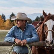 kevin costner in yellowstone next to a horse leaning on a fence with rope in hands wearing a faded blue denim shirt and beige cowboy hat