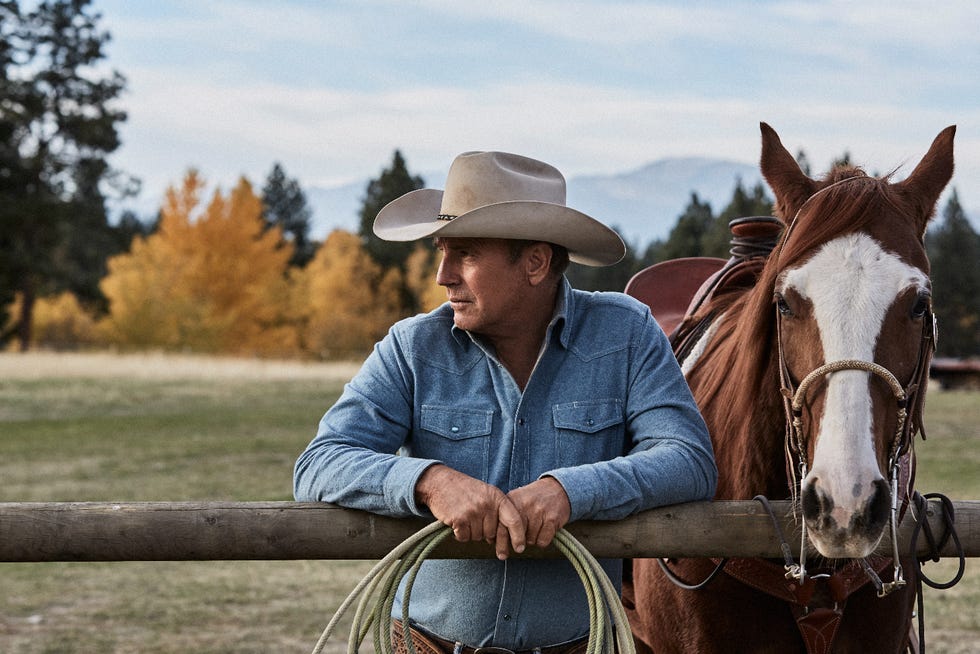 kevin costner in yellowstone next to a horse leaning on a fence with rope in hands wearing a faded blue denim shirt and beige cowboy hat