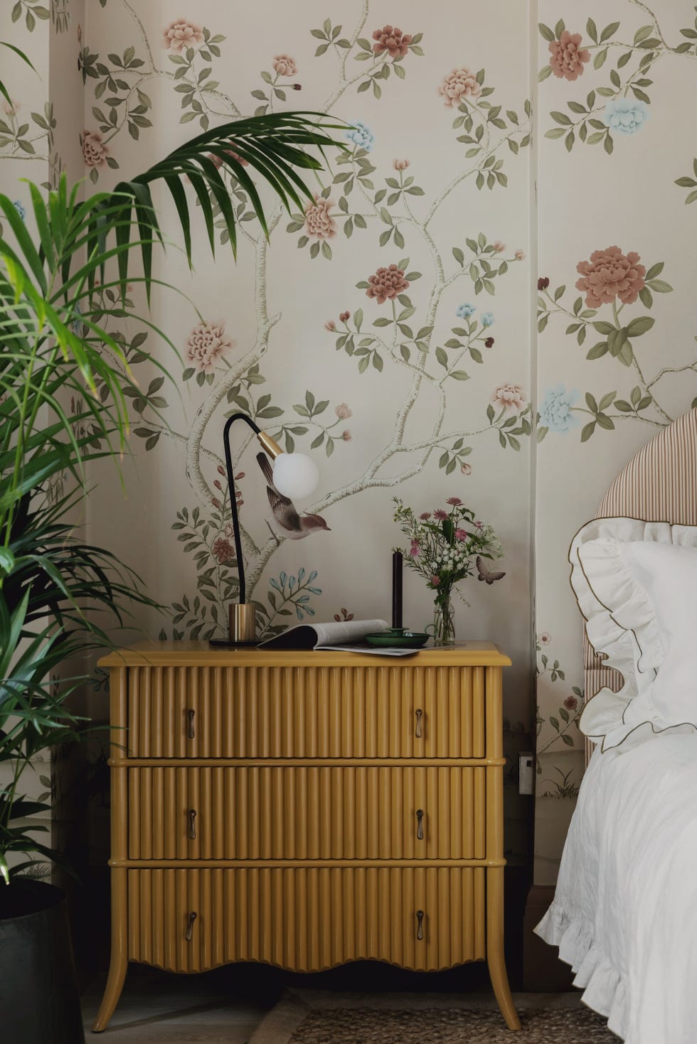 nightstand beside a bed with a decorative lamp and flowers set against floral wallpaper
