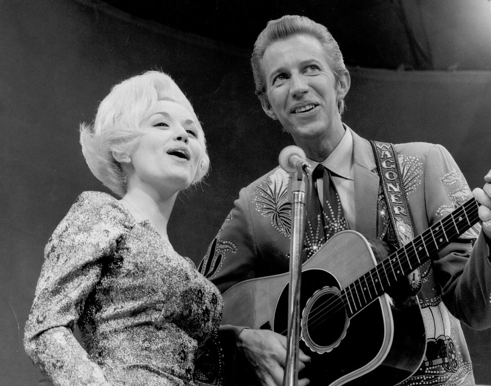 a black and white photo of dolly parton, wearing a sparkling dress, and porter wagoner, wearing a suit and holding a guitar, as they both sing into a microphone