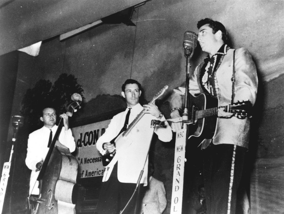 marshall grant, luther perkins, and johnny cash perform on a stage, each wears a suit jacket, tie and dark pants, cash sings into a microphone on a stand