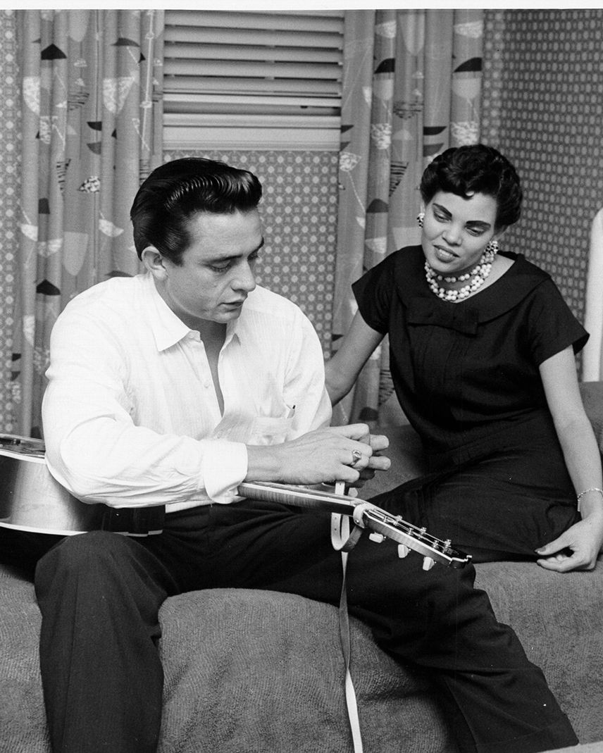 johnny cash and vivian liberto sit on a twin bed, he works on a guitar strap as his instrument lays in his lap, she looks on