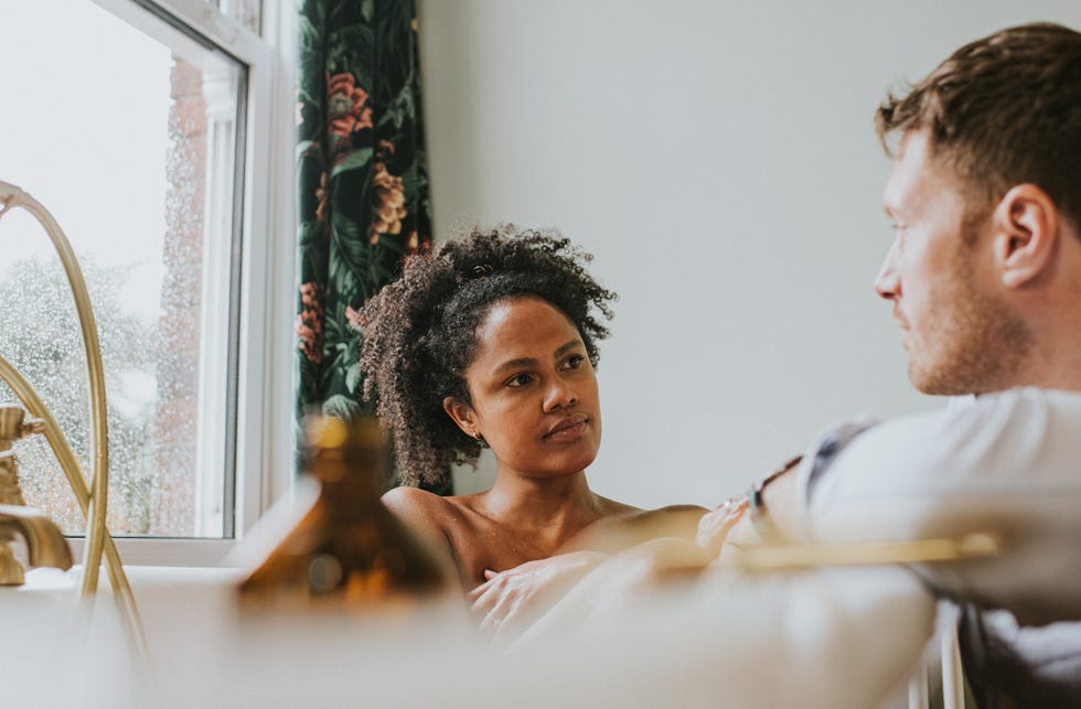 a couple have a conversation as the woman bathes and her partner leans on the side of the tub