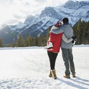 winter date ideas   couple with ice skates hugging below snowy mountain