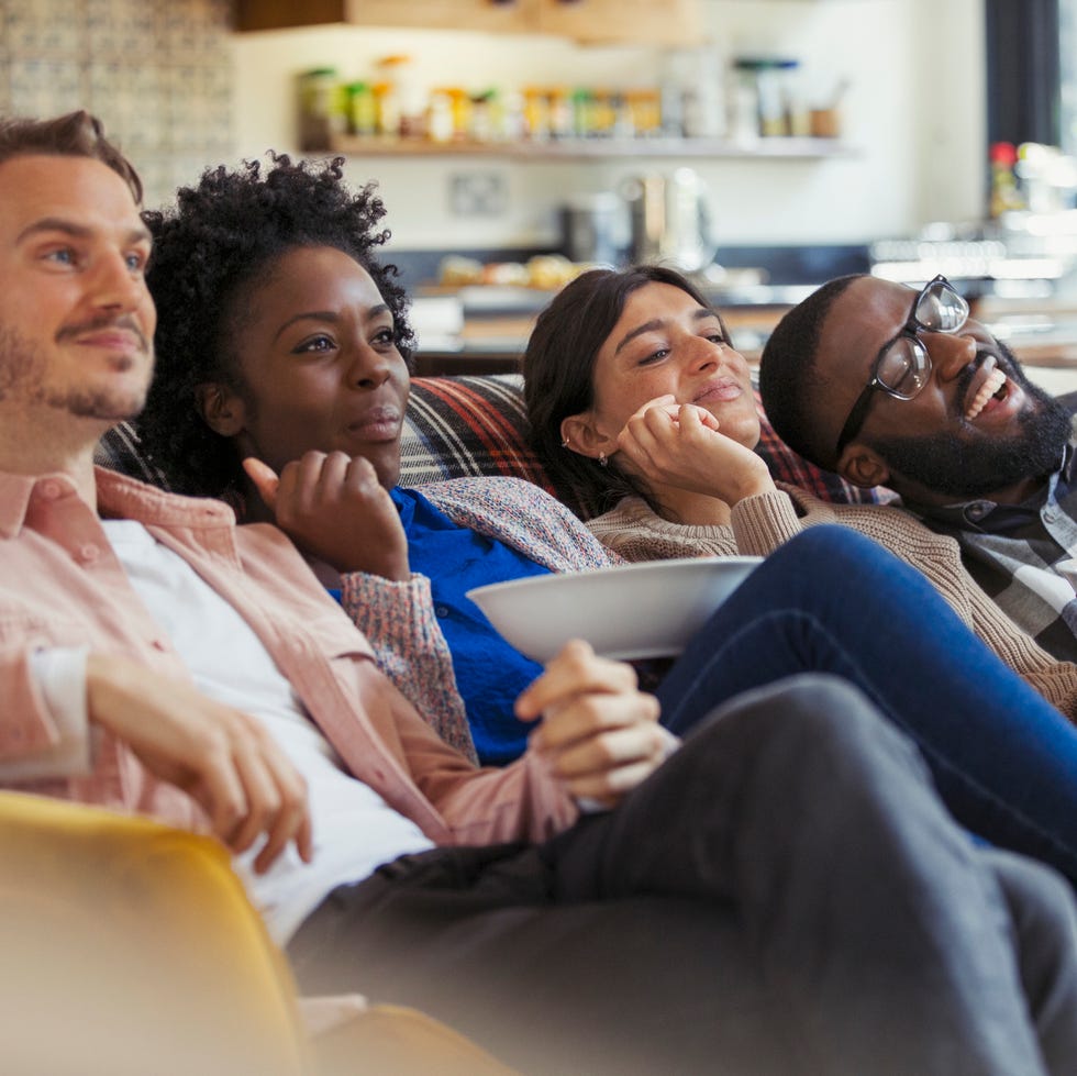 a white man, black woman, white woman, and white man watching tv and eating popcorn on living room sofa