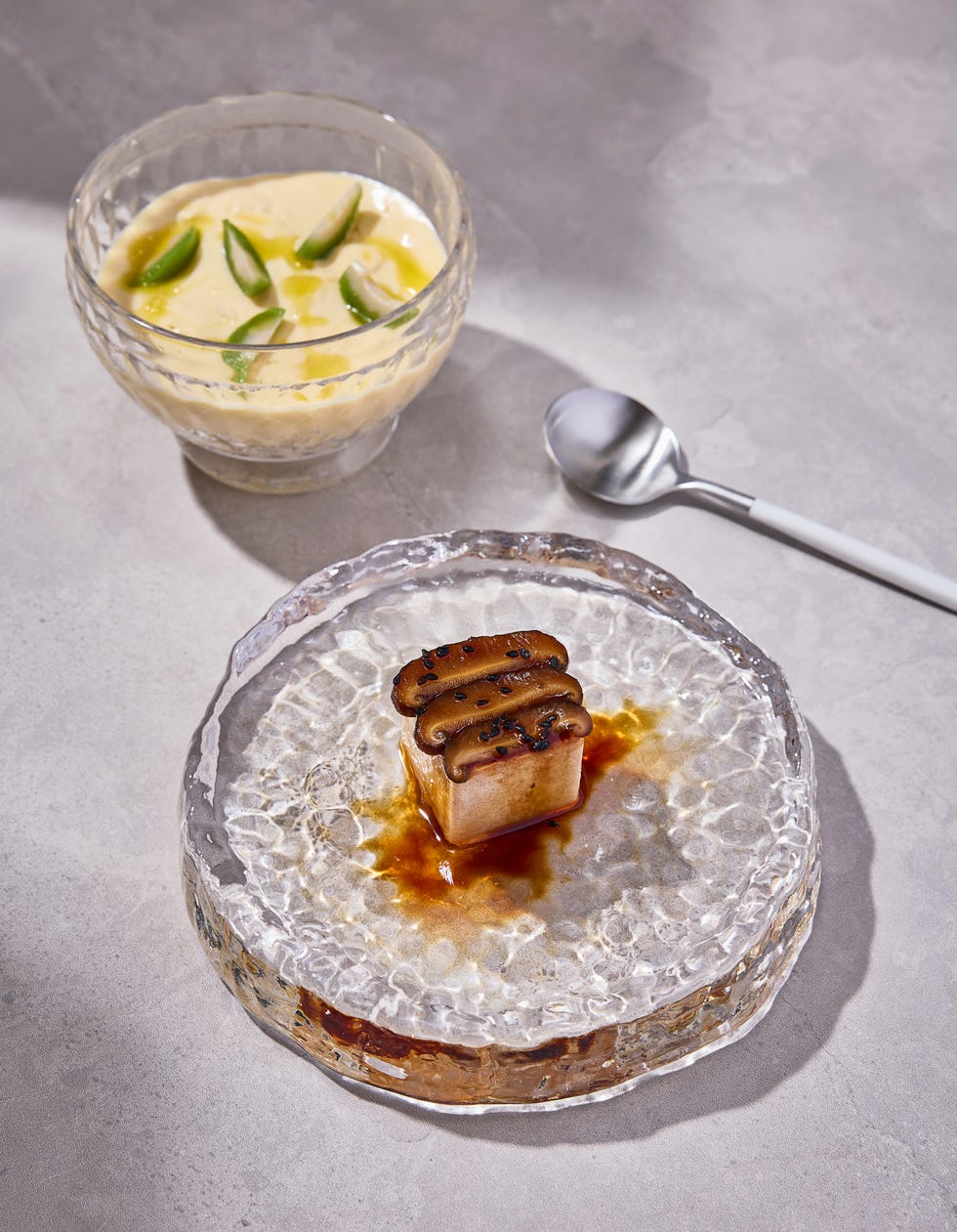 a dessert plate featuring a layered rectangular dessert with a glossy glaze and adorned with decorative elements alongside a bowl of custard topped with green accents