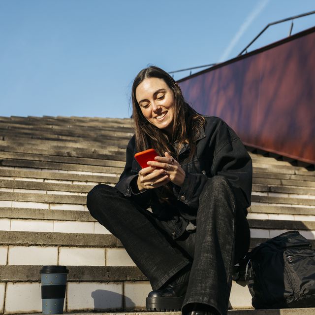 brunette young woman sitting on a public park stairs and texting on her red smartphone