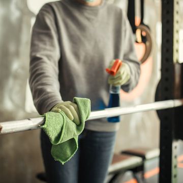 mature woman cleaning and disinfecting equipment in gym without people she is using microfiber cloth and antiseptic liquid spray and she wears protective cap, mask and gloves disinfection and hygiene in gym is very important to stop spreading virus infections photo taken during corona virus outbreak