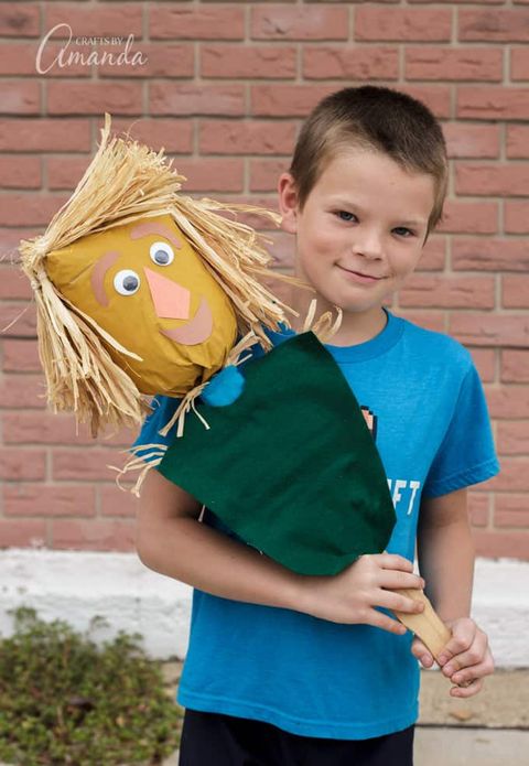 smiling little boy holding homemade scarecrow puppet on a stick made from a paper bag with straw hair and googly eyes