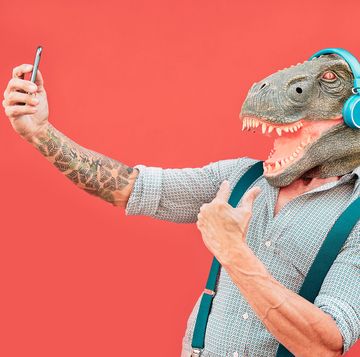 crazy senior man wearing t rex mask and taking selfie with mobile smartphone hipster older male having fun listening music and dancing outdoor absurd, funny and surreal concepts