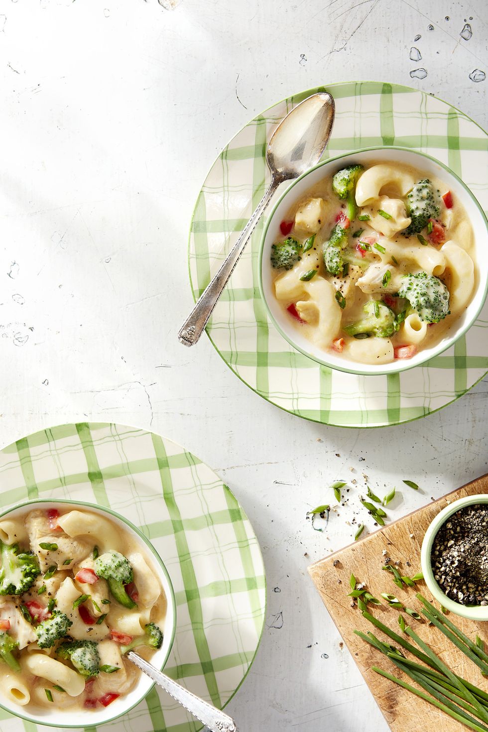 2 bowls of creamy macaroni pasta soup with broccoli, set on summery green and white plaid patterned plates