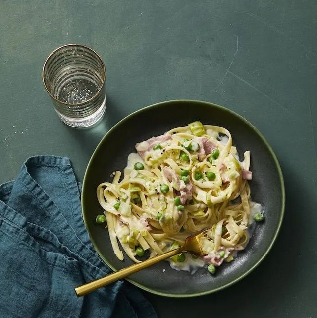 a dark gray bowl of creamy fettuccine with leeks and ham with a gold fork, dark blue napkin and a glass of sparkling water