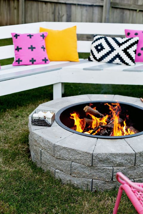 diy circular stone outdoor fireplace in grass next to white garden bench and pink, yellow, black, and white pillows
