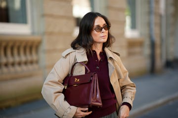 una mujer viste un jersey tipo polo de punto burdeos con tres botones, cuello y dobladillo acanalado; un clásico abrigo largo beige de herno, con solapa de muesca, solapa cortavientos y puños con cinturón; un bolso estructurado de cuero burdeos brillante modelo kelly de hermès con herrajes en tono dorado; unas gafas de sol ovaladas de carey de miu miu; su cabello castaño está peinado liso, hasta los hombros, con raya en el medio.