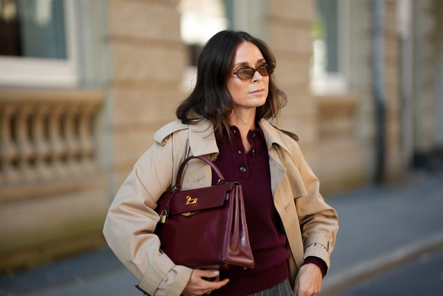 una mujer viste un jersey tipo polo de punto burdeos con tres botones, cuello y dobladillo acanalado; un clásico abrigo largo beige de herno, con solapa de muesca, solapa cortavientos y puños con cinturón; un bolso estructurado de cuero burdeos brillante modelo kelly de hermès con herrajes en tono dorado; unas gafas de sol ovaladas de carey de miu miu; su cabello castaño está peinado liso, hasta los hombros, con raya en el medio.