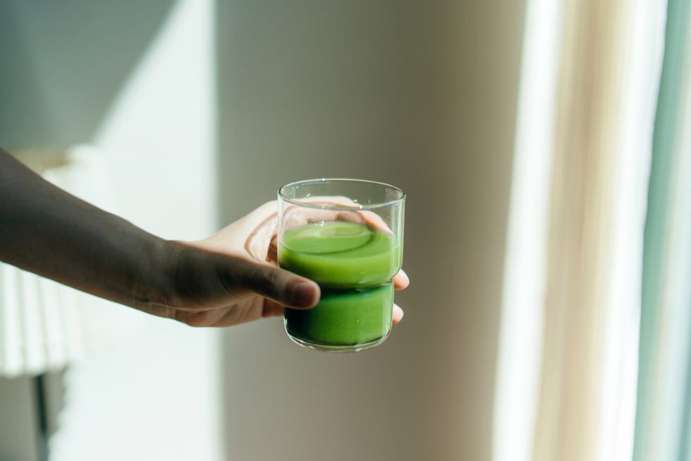 cropped hand of woman holding a glass of fresh green fruit and vegetable smoothie against white background with natural sunlight healthy superfood green colour, detox diet and healthy eating concept