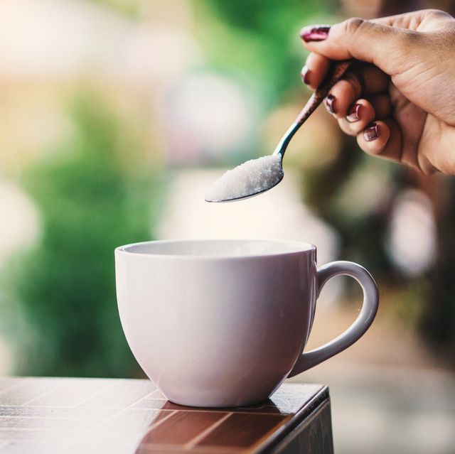 cropped hand of woman holding spoon with sugar over coffee cup on table