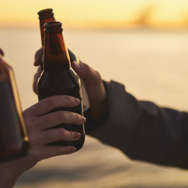 cropped image of friends toasting beer bottles against sea during sunset