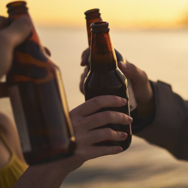 cropped image of friends toasting beer bottles against sea during sunset
