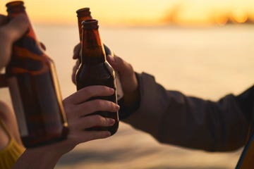 cropped image of friends toasting beer bottles against sea during sunset