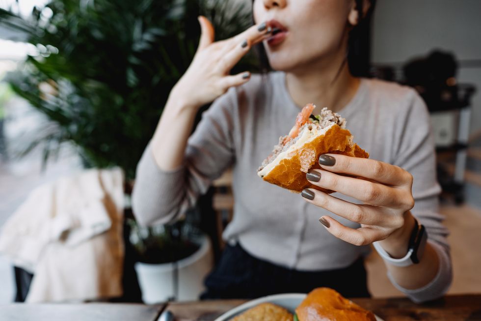 cropped shot, mid section of young asian woman eating freshly made delicious cheeseburger in a cafe, licking her fingers enjoying her lunch people and food concept