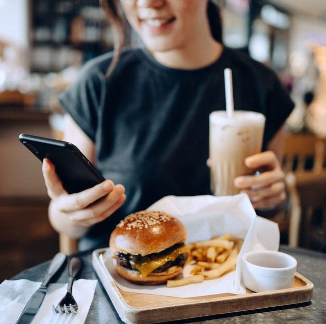 cropped shot of smiling young asian woman using smartphone while enjoying her lunch, cheeseburger with french fries and iced coffee in a stylish cafe people, food, lifestyle and technology concept