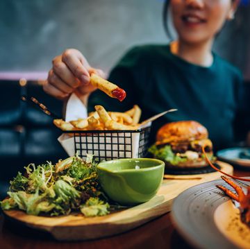 cropped shot of young asian woman eating freshly made delicious burger, dipping fries in ketchup on the dining table enjoying a feast in the restaurant people, food and lifestyle
