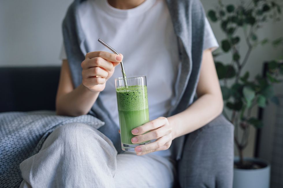 cropped shot of young asian woman sitting on the sofa at home, drinking a glass of fresh green detox juice with reusable metal straw healthy eating, green colour and detox diet concept