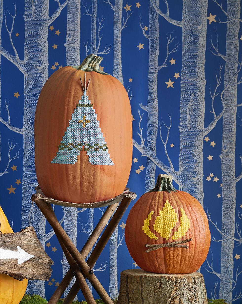 teepee and flame designs cross stitched on pumpkins, atop rustic wood stools with wallpaper backdrop of forest at night
