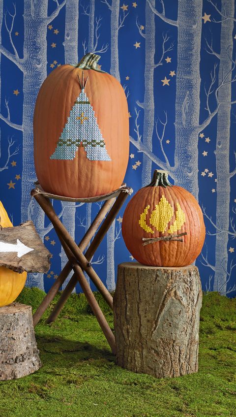 teepee and flame designs cross stitched on pumpkins, atop rustic wood stools with wallpaper backdrop of forest at night
