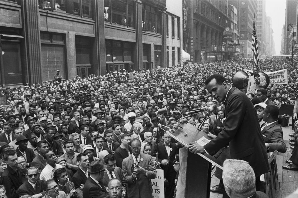 harry belafonte sings into a microphone while on stage, in front of him a city street is packed full of demonstrators