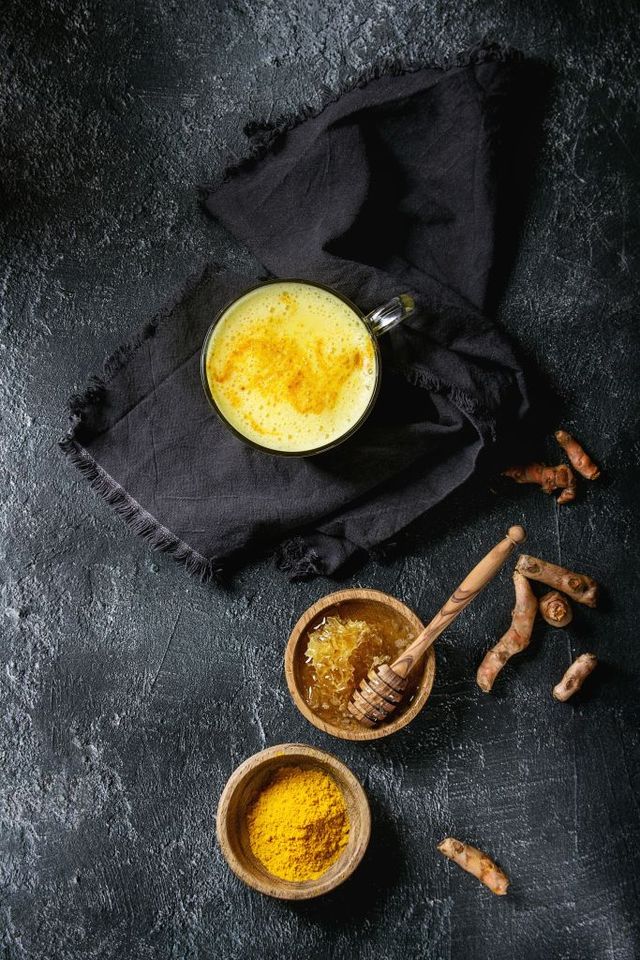 Cup of ayurvedic drink golden milk turmeric latte with curcuma powder and ingredients above over black texture background Top view, space