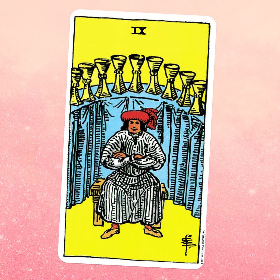 the tarot card the nine of cups, showing a person in a white robe and red hat sitting with their arms crossed in front of a tall table on which nine gold cups are placed