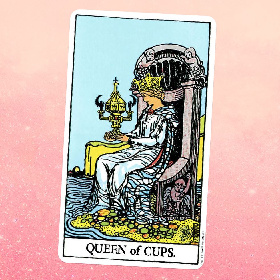 the tarot card the queen of cups, showing a person in a long white robe and a crown sitting on a throne on a seashore and holding up a giant goblet