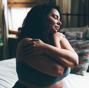 curly haired overweight young woman in blue top and shorts with satisfaction on face accepts curvy body shape in stylish bedroom