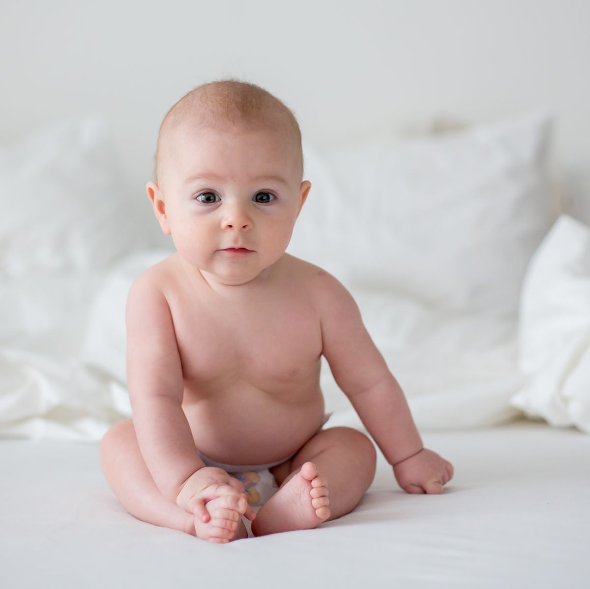 Cute little baby boy in diaper, smiling at camera in white bedroom, cute toddler sitting on bed, smiling
