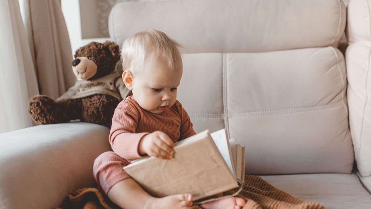 cute smiling little baby reading book sitting on sofa with teddy bear toy and bunny rabbit at home