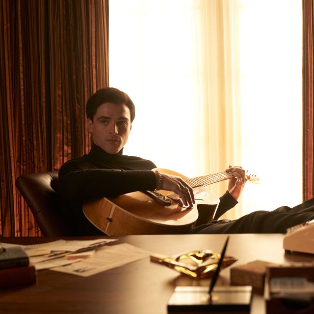 jacob elordi sitting with his feet propped up on a desk and holding a guitar