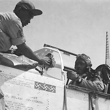 clarence lester sits in the open cockpit of a world war ii fighter plane, while an engineer works on the front of the plane